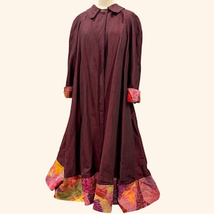 Puede incluir: Un abrigo largo burdeos con cuello redondo y costuras decorativas. Los puños y el dobladillo presentan un diseño de patchwork con colores vibrantes como el rojo, el naranja y el amarillo. El abrigo se exhibe en un maniquí.