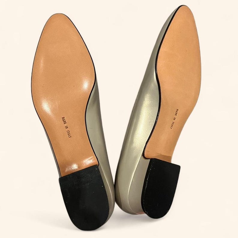May include: A pair of metallic silver-colored low-heeled shoes. The soles are light brown with the words "MADE IN ITALY" printed on them. The heels are black and the shoes have a pointed toe.
