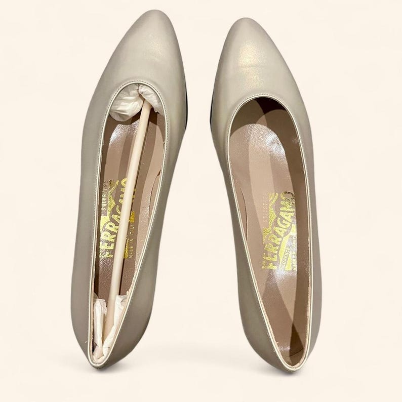 May include: A pair of light gray pointed-toe flats. The shoes have a metallic sheen and are displayed with the original packaging inside. The interior of the shoe has a gold logo. The shoes are a classic style.