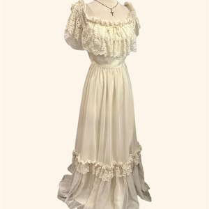 May include: A floor-length cream-colored dress featuring a ruffled lace neckline and short puff sleeves. The bodice is fitted, transitioning into a flowing skirt with a horizontal lace-trimmed ruffle near the hem. A thin necklace with a cross pendant is visible.