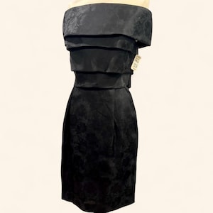 Puede incluir: Vestido negro con hombros descubiertos, corpiño en capas y falda hasta la rodilla. El vestido tiene un sutil estampado floral y una etiqueta adjunta. El vestido se exhibe en un maniquí.