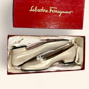 May include: A pair of cream-colored Salvatore Ferragamo pumps in their original red box. The shoes have a low heel and a rounded toe. The box has the brand name in gold lettering on the lid.