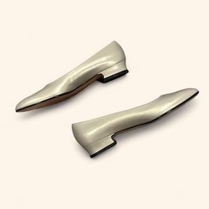 May include: A pair of ivory-colored women's flats with a low heel. The shoes have a pointed toe and a smooth, shiny finish. The heels are black, and the soles are brown.