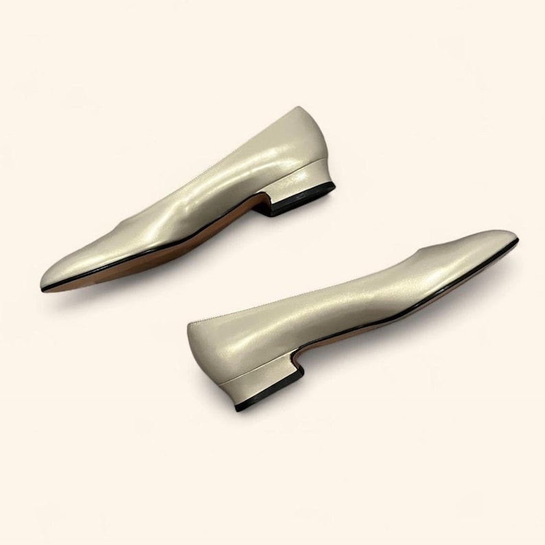 May include: A pair of ivory-colored women's flats with a low heel. The shoes have a pointed toe and a smooth, shiny finish. The heels are black, and the soles are brown.