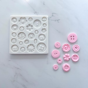 May include: A white square silicone mold featuring various circular button shapes with different patterns and hole configurations, accompanied by ten individual light pink buttons cast from the mold, displaying floral, smooth, and textured designs.