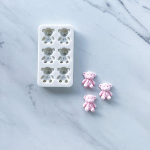 May include: A white rectangular silicone mold featuring six recessed cavities shaped like teddy bears, accompanied by three small, light pink, textured teddy bear figures that have been cast from the mold.