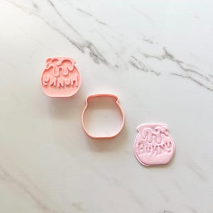 May include: Three light pink plastic cookie cutters shaped like honey pots. Two of the cutters feature an embossed design of dripping honey and the word HUNNY, while the center cutter is a simple outline of a honey pot without any interior details.