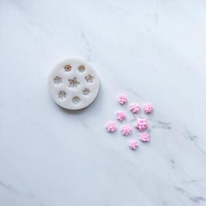 May include: A circular white silicone mold featuring eight distinct cavities shaped like various succulent plants, accompanied by nine small, light pink, molded succulent plant figures arranged in a cluster to the right of the mold.