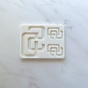 May include: A rectangular white silicone mold featuring recessed channels in a geometric maze-like pattern. The design includes one large, complex interlocking shape on the left side and two smaller, identical interlocking shapes on the right side.