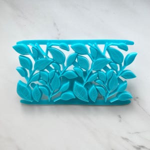 May include: A bright turquoise plastic mold featuring a repeating pattern of detailed leaves. The rectangular tool has a horizontal bar at the top and bottom, with intricate leaf shapes connected by thin stems throughout the center section.