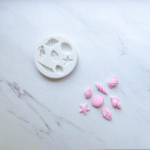 May include: A round white silicone mold featuring recessed shapes of various seashells and a starfish, accompanied by seven small, light pink molded seashell and starfish figures arranged in a cluster to the lower right of the mold.