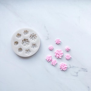 May include: A round white silicone mold featuring eight distinct floral and succulent cavities, accompanied by eight small, light pink molded flower shapes that correspond to the designs found within the mold's circular layout.
