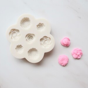 May include: A white silicone mold featuring seven distinct floral cavities in various sizes and shapes, designed for creating detailed flower-shaped castings. Three small, pink, finished flower-shaped pieces are positioned to the right of the mold.