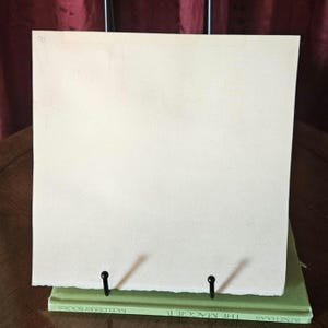 Pu&ograve; includere: Un foglio di carta bianco sporco vuoto &egrave; esposto su un supporto di metallo nero. Il supporto &egrave; posizionato sopra un libro verde. La carta ha una superficie leggermente strutturata ed &egrave; tenuta in posizione da due supporti di metallo nero.