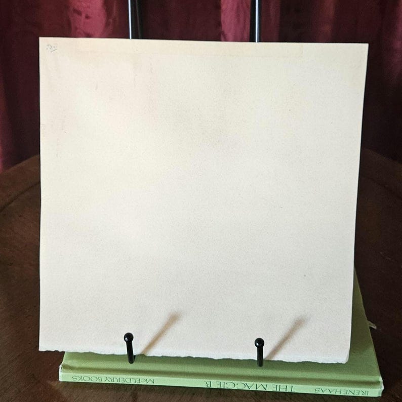 Pu&ograve; includere: Un foglio di carta bianco sporco vuoto &egrave; esposto su un supporto di metallo nero. Il supporto &egrave; posizionato sopra un libro verde. La carta ha una superficie leggermente strutturata ed &egrave; tenuta in posizione da due supporti di metallo nero.