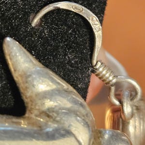 Puede incluir: Primer plano de un pendiente de plata con un gancho curvo y un elemento decorativo. El gancho tiene una peque&ntilde;a marca. El pendiente est&aacute; sobre un fondo negro, con un fondo naranja borroso.