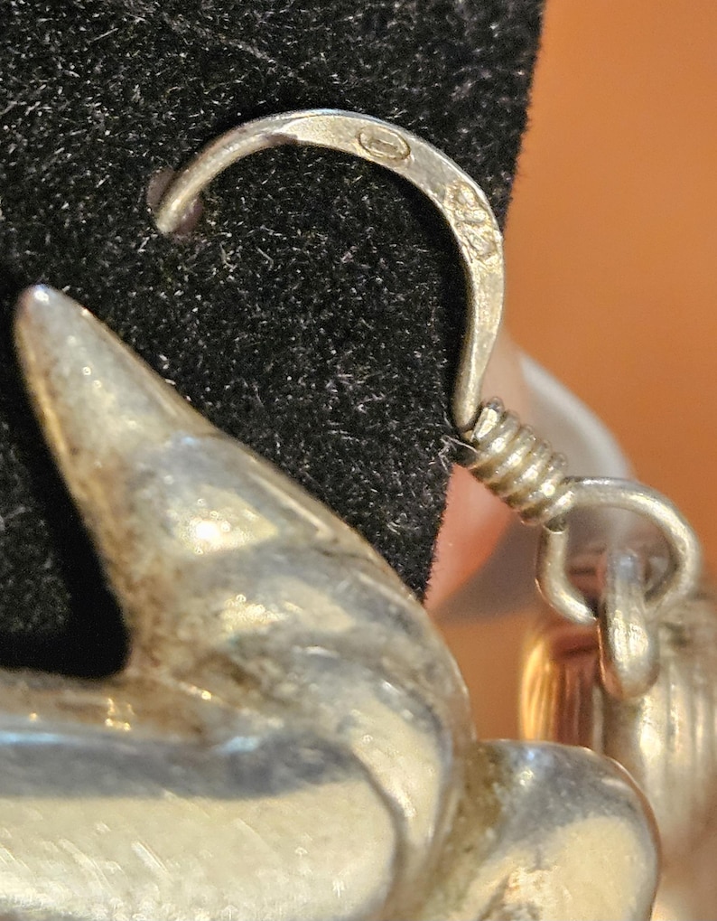 Puede incluir: Primer plano de un pendiente de plata con un gancho curvo y un elemento decorativo. El gancho tiene una peque&ntilde;a marca. El pendiente est&aacute; sobre un fondo negro, con un fondo naranja borroso.