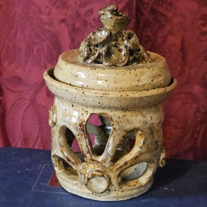 May include: A speckled beige ceramic jar with a lid and decorative cutouts. The jar has a round shape with a lid that features a floral design. The cutouts are in a leaf-like pattern. The jar is sitting on a blue surface.