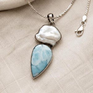 May include: A silver necklace featuring a pendant with a white baroque pearl positioned above a teardrop-shaped light blue Larimar stone. The pendant is attached to a delicate silver box chain with a lobster claw clasp closure.