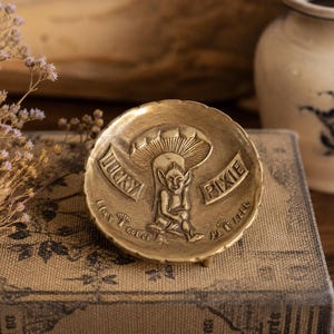 May include: A round brass dish featuring a central embossed design of a pixie sitting under a mushroom. The text "LUCKY PIXIE" is engraved on banners flanking the figure, with additional small text below the pixie. The dish has a scalloped edge.