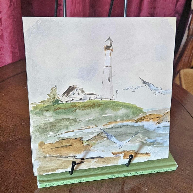 Pu&ograve; includere: Dipinto ad acquerello di un faro su una collina erbosa con un edificio bianco. Due gabbiani volano nel cielo. Il dipinto &egrave; esposto su un leggio verde. Il titolo del libro &egrave; "The Maggie B."