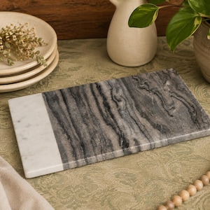 May include: A rectangular serving board made of natural stone, featuring a distinct color-blocked design. One side is solid white marble, while the larger portion displays a dark gray and black swirling marble pattern with a smooth, polished finish.
