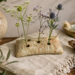 May include: A rectangular ceramic flower frog with a mottled beige and tan glaze featuring five circular openings. The piece holds several delicate stems, including white lace flowers, small purple blossoms, and a single blue thistle head.