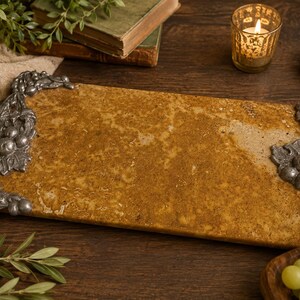 May include: A rectangular serving tray featuring a textured, golden-brown stone surface. The tray is accented on two opposite ends with ornate, metallic silver-toned handles shaped like clusters of grapes, leaves, and vines in a detailed relief.