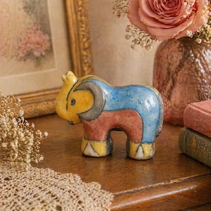 May include: A colorful ceramic elephant figurine. The elephant is painted with yellow, blue, red, and gray sections. The figurine is displayed on a wooden surface, with a framed picture and a vase of flowers in the background.