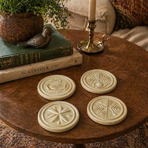 May include: Four beige ceramic coasters with embossed designs sit on a wooden table. The designs include a swan, a star, and two different leaf patterns. A small bronze bird and a stack of books are also on the table.