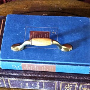May include: A brass and wood drawer pull rests on a blue book titled "The Adventures of Sherlock Holmes". The hardware features a light wood center and curved brass ends. The book is stacked on top of other antique books.