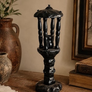May include: A black wooden candle holder featuring a textured, carved base and a central pillar. The top section consists of a circular roof supported by four vertical spindles, surrounding a small central cup designed to hold a single candle.