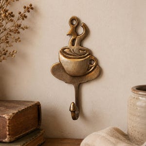 May include: A wall-mounted metal hook with a bronze-toned finish, featuring a decorative design of a coffee cup on a saucer with stylized steam rising from the top. The hook extends downward from the base of the saucer, providing a functional hanger.