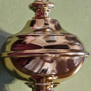 May include: A gold-colored decorative finial with a bulbous body and a slender, tiered top. The finial has a threaded base and is likely for a lamp or furniture. The surface reflects light.