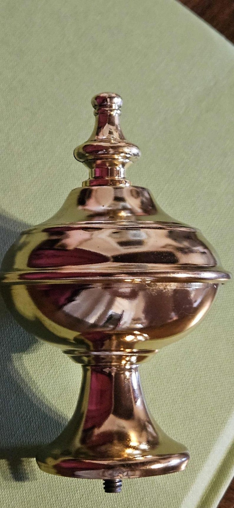 May include: A gold-colored decorative finial with a bulbous body and a slender, tiered top. The finial has a threaded base and is likely for a lamp or furniture. The surface reflects light.
