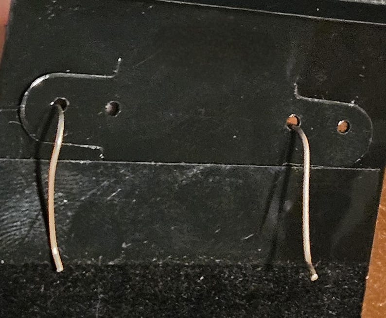 Puede incluir: Un par de pendientes delicados y finos, de color plateado, colgando de una tarjeta de exhibici&oacute;n de pendientes negra. Los pendientes son sencillos, con un dise&ntilde;o curvo similar a un alambre. La tarjeta tiene dos agujeros para colgar los pendientes.