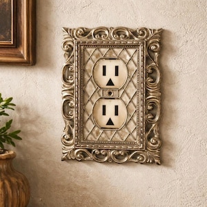 May include: Ornate, antique-style electrical outlet cover with a decorative frame. The cover features two standard outlets with black inserts and a diamond-patterned background. The frame has a silver finish and intricate scrollwork.