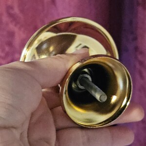 May include: Close-up of a gold-colored furniture foot with a threaded bolt. The foot has a rounded, bell-like shape and a shiny finish. The bolt is silver and extends from the center.
