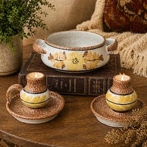 May include: A ceramic bowl with handles and two matching candle holders. The bowl and candle holders are white with yellow and brown floral designs. The candle holders have lit candles inside.