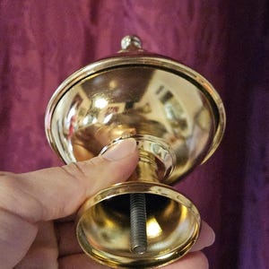 May include: A gold-colored, decorative finial with a rounded top and a small, spherical detail. The finial has a flared base and a threaded rod extending from the bottom. The object is held in a hand.