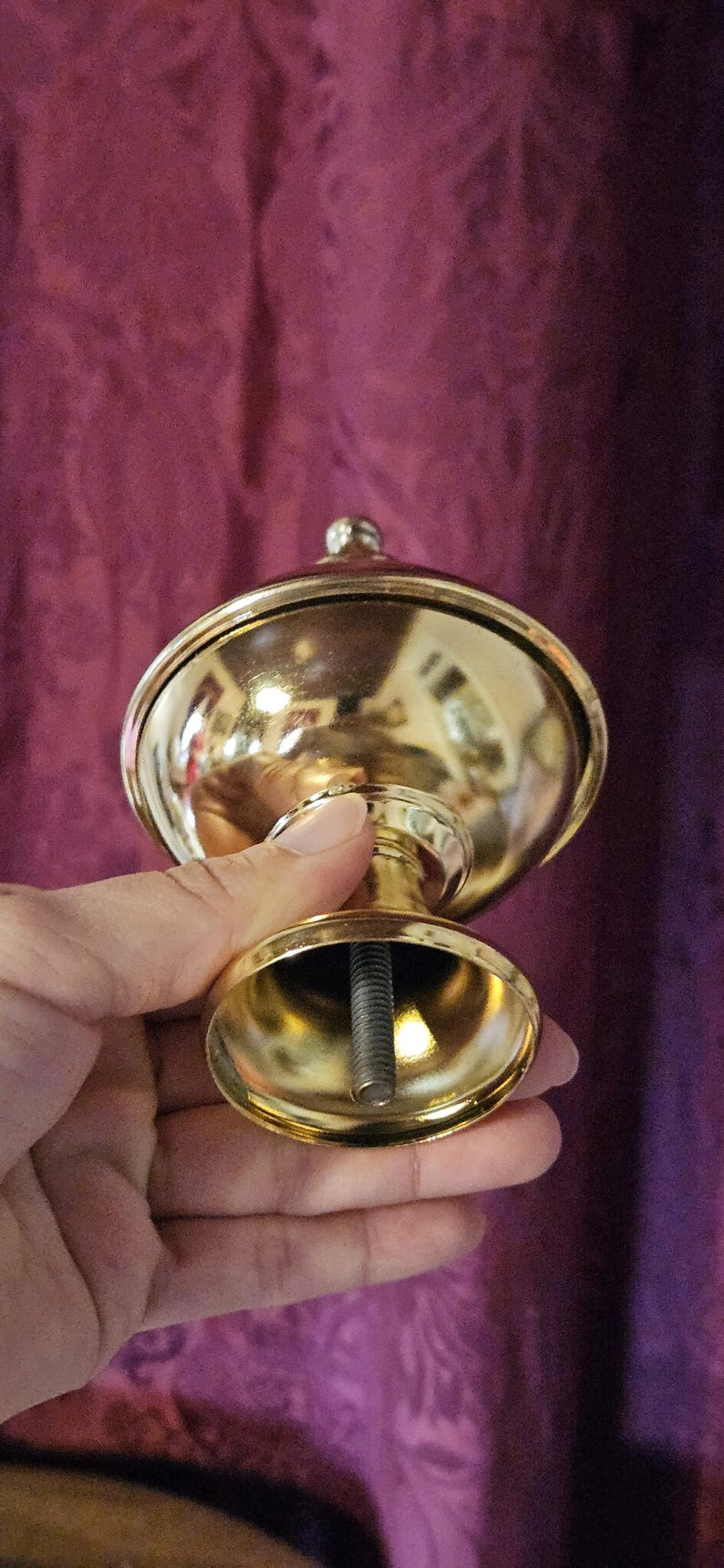 May include: A gold-colored, decorative finial with a rounded top and a small, spherical detail. The finial has a flared base and a threaded rod extending from the bottom. The object is held in a hand.
