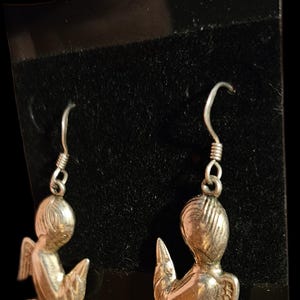 Puede incluir: Un par de pendientes de &aacute;ngel plateados. Cada pendiente presenta una peque&ntilde;a y detallada figura de &aacute;ngel con alas, arrodillada con las manos juntas en oraci&oacute;n. Los pendientes tienen un dise&ntilde;o de gancho simple para orejas perforadas, sobre un fondo oscuro.