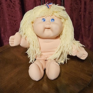May include: A vintage Cabbage Patch Kid doll with a soft, fabric body and a vinyl head. The doll has blonde yarn hair, blue eyes, and a small ribbon on top of its head. The doll is sitting on a brown surface.
