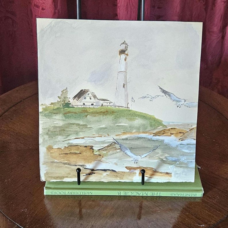 Pu&ograve; includere: Dipinto ad acquerello di un faro su una collina verde con un edificio bianco. L'opera presenta gabbiani in volo e una costa rocciosa. Il dipinto &egrave; esposto su un libro verde con il titolo "THE MAGGIE B."