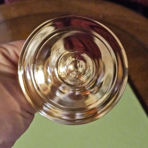 May include: A gold-colored, ornate chalice or goblet, viewed from above. The cup has a wide, circular base and a detailed, ridged design. The surface reflects light, creating a shiny appearance. The background is a muted green and purple.