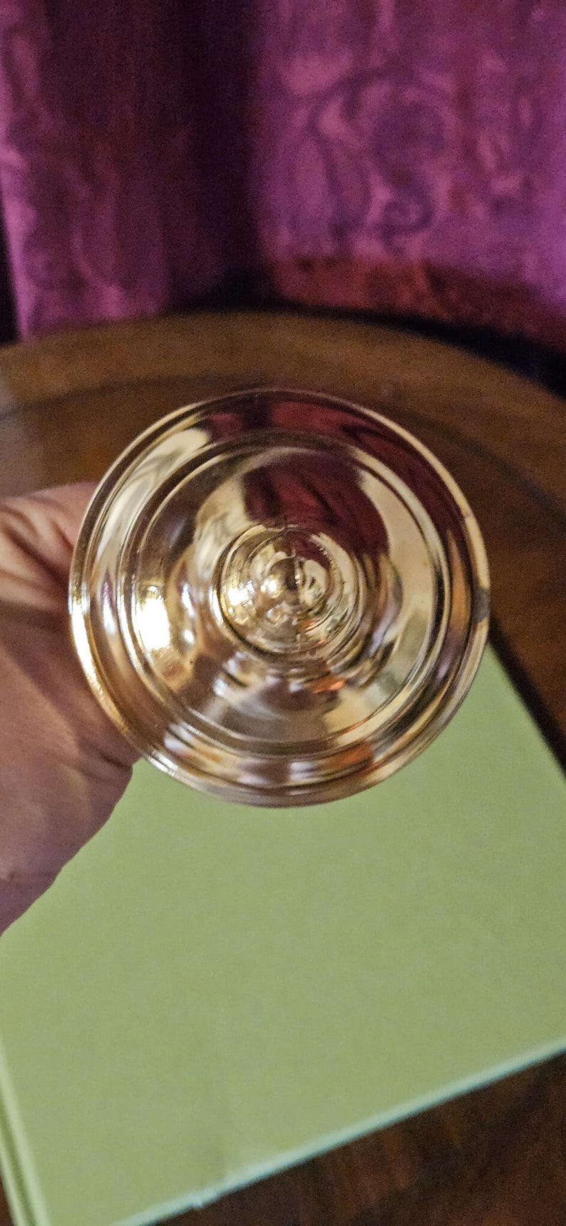May include: A gold-colored, ornate chalice or goblet, viewed from above. The cup has a wide, circular base and a detailed, ridged design. The surface reflects light, creating a shiny appearance. The background is a muted green and purple.