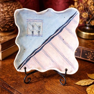 May include: A square ceramic plate with a wavy edge, resting on a black metal stand. The plate features a light blue background with a diagonal section of pink stripes and a small illustration of figures.