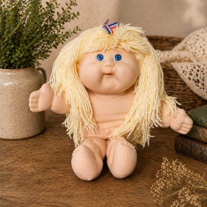 May include: A vintage doll with a soft, flesh-toned body, blue eyes, and blonde yarn hair. The doll has a small bow with red, white, and blue accents. The doll is sitting on a wooden surface.