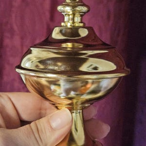 May include: A gold-colored, ornate decorative container with a lid, held by a hand. The container has a rounded body and a pedestal base. The lid is topped with a decorative finial. The background is a deep purple.