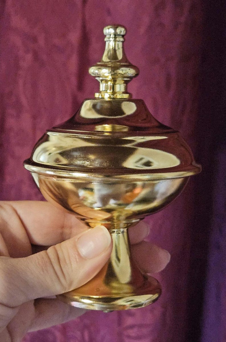 May include: A gold-colored, ornate decorative container with a lid, held by a hand. The container has a rounded body and a pedestal base. The lid is topped with a decorative finial. The background is a deep purple.